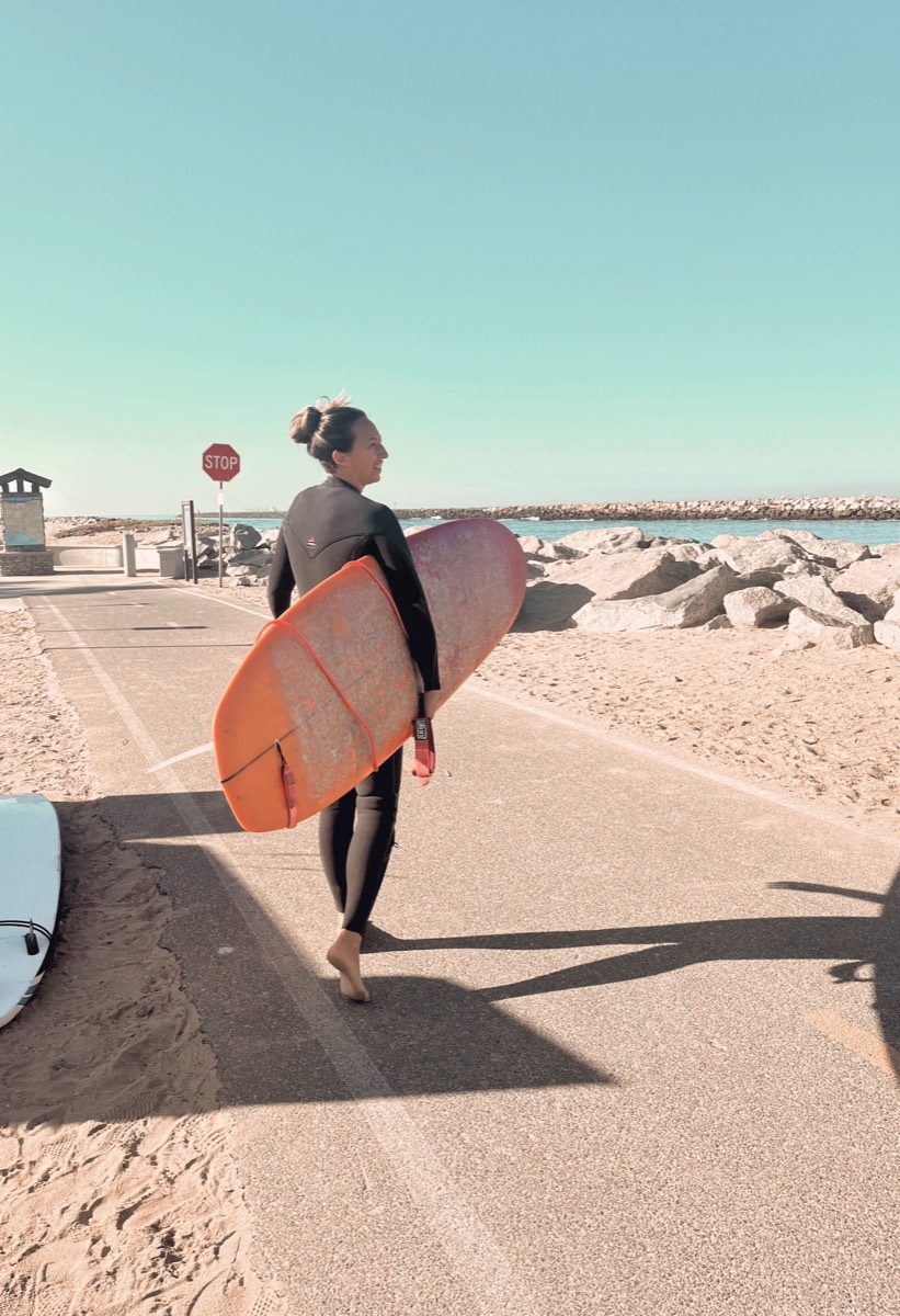 Crystal Ortiz carrying her orange surfboard along the beach path in Long Beach